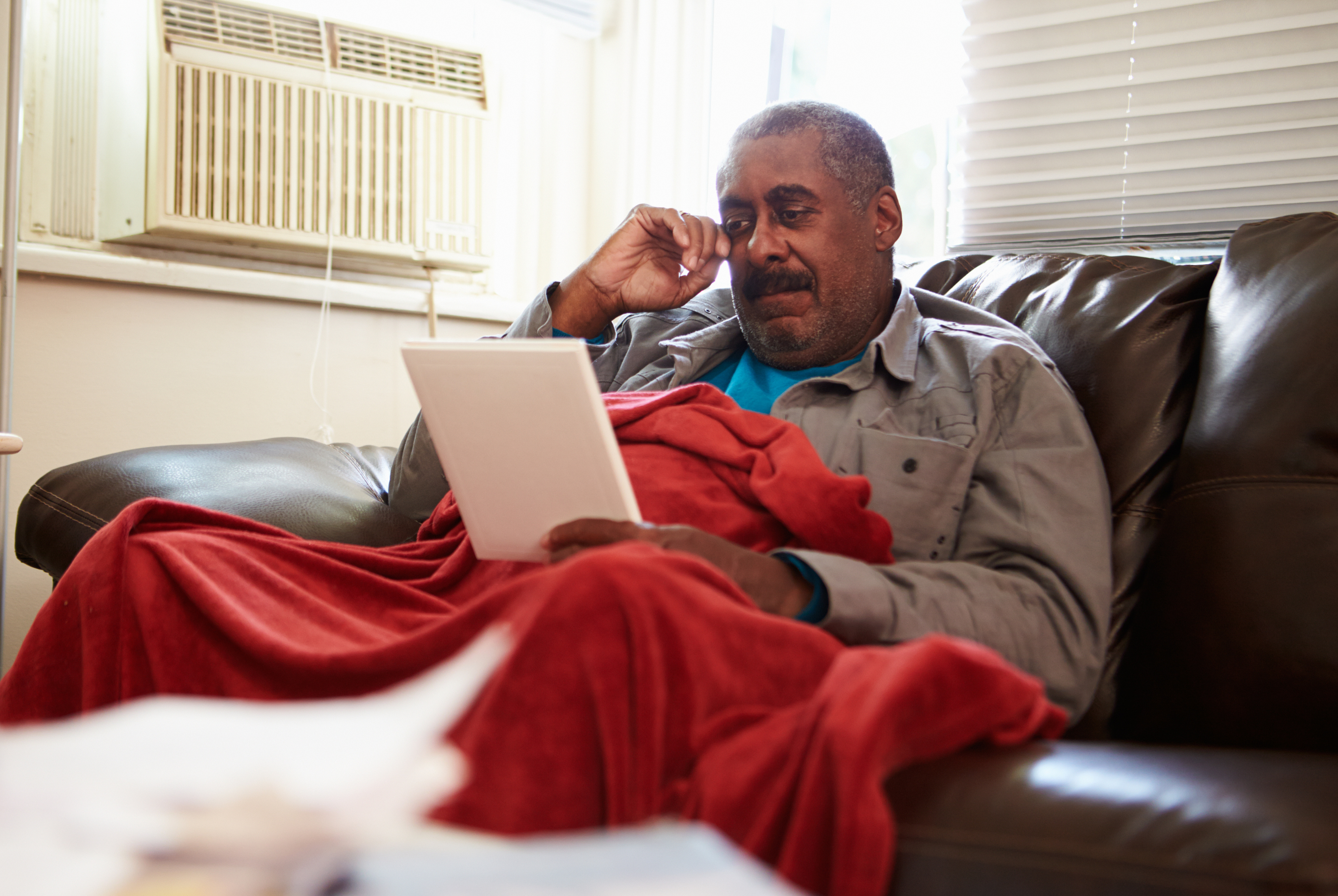 A man sitting on a sofa with a blanket trying to keep warm