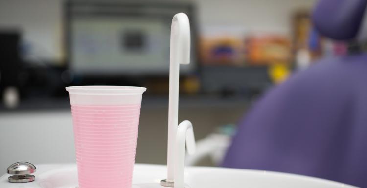 Image of a dentist's room showing tap and cup full of pink rinsing liquid with dentist's chair in background.