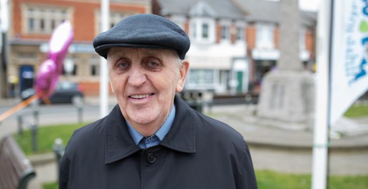 An older man wearing a hat standing out doors and smiling at the camera