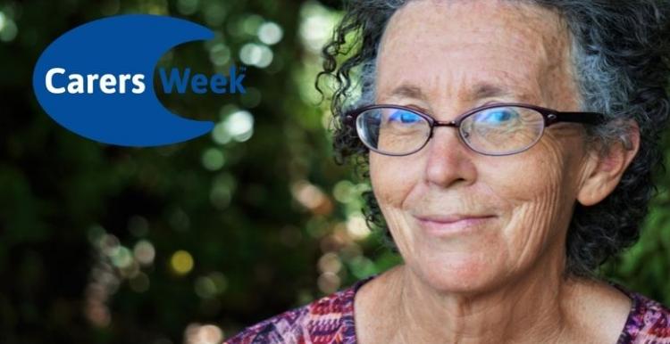 A woman wearing glasses is looking into the camera lens and smiling with a closed mouth. Logo in the corner says Carers Week.