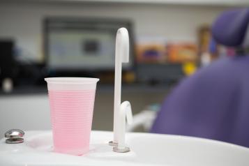 Image of a dentist's room showing tap and cup full of pink rinsing liquid with dentist's chair in background.