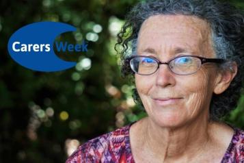 A woman wearing glasses is looking into the camera lens and smiling with a closed mouth. Logo in the corner says Carers Week.