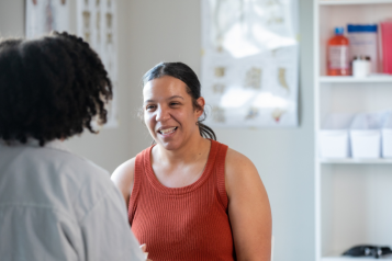 A lady talking to a person in white with medical bottles on a shelf in the background.