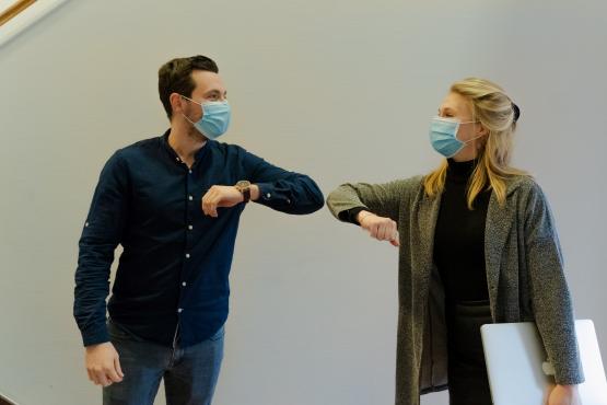 A man and a woman in facemasks look at each other whilst bumping elbows. 